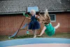 Two young girls jumping on a trampoline with a slide and swing set in the back ground.